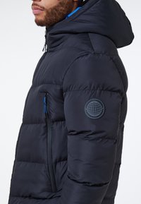 Uomo che indossa una giacca a vento nera con cappuccio, tasca laterale con zip e linguette blu, e logo circolare del globo sulla manica superiore sinistra.