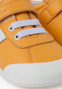 Sneaker in pelle gialla con suola in gomma bianca, dotato di lacci elastici doppi e una fascia con velcro per una vestibilità sicura. Texture liscia.