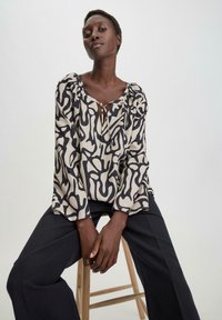 Blusa leggera in crema con motivi astratti neri, scollo a barco e maniche a palloncino. Abbinata a pantaloni larghi scuri.