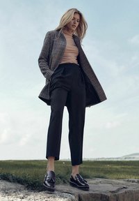 Woolowy płaszcz w kratę houndstooth na beżowym golfie, wysokie czarne spodnie i czarne loafersy z frędzlami. Ustawienie na świeżym powietrzu.