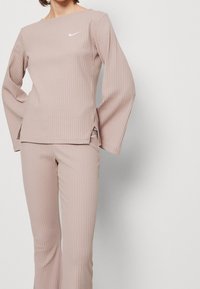 Světle taupe pruhovaný top s dlouhým rukávem a kulatým výstřihem, doplněný o kalhoty se stejným pruhováním. Na hrudi se nachází malé bílé logo.