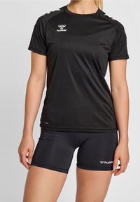 Musta lyhythihainen urheilupaita, jossa on pyöreä kaula-aukko. Sisältää valkoisen logon ja olkapään korostuksia. Yhdistettynä samanlaisiin mustiin shortsit.