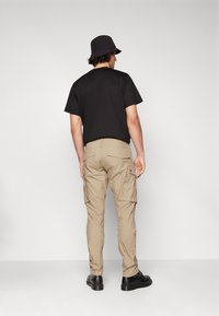 Beige Cargo-Hosen mit mehreren Seitentaschen, getragen mit einem schwarzen T-Shirt und einem schwarzen Bucket Hat. Die Hose hat eine taillierte Passform und sichtbare Nähte.