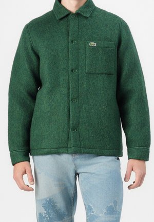 Mężczyzna ubrany w zieloną, zapinaną na guziki wełnianą kurtkę z kieszenią na piersi i logo, w połączeniu z jasnoniebieskimi dżinsami z wybielonymi wzorami.