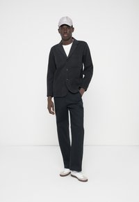 Juodas audinio švarkas su įpjautomis atlapais, priekinėmis kišenėmis, derinamas su atitinkančiomis kelnėmis ir baltais sportbačiais. Šviesiai pilka kepurė užbaigia aprangą.