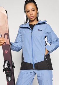 Ανοιχτό μπλε, αδιάβροχο μπουφάν με μαύρες λεπτομέρειες, πλήρης φερμουάρ, ρυθμιζόμενη κουκούλα και ελαστικές μανσέτες. Το μοντέλο κρατάει ένα σκι με σκούρα μωβ γραφικά.