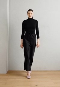 Haut à col roulé noir à manches longues associé à une jupe noire longue, avec une texture lisse et des chaussures roses sur un sol en bois.