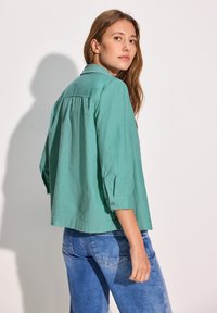 Camisa de manga larga verde claro con cuello y puños abotonados, de corte holgado y con detalle de pliegue en la parte posterior, combinada con jeans de mezclilla azul.
