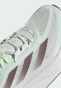 Scarpa atletica verde chiaro con tomaia in mesh, accenti testurizzati, strisce viola e suola bianca. Presenta una linguetta e lacci imbottiti.