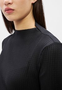 Žena s rovnými tmavými vlasmi, oblečená v čiernom rebrovanom tričku s dlhým rukávom a vysokým výstrihom, na jednoduchom svetlom pozadí.