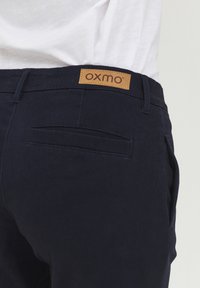 Marineblå bomullsbukser med en lærmerke merket "OXMO" på linningen. Har baklommer og en jevn tekstur.