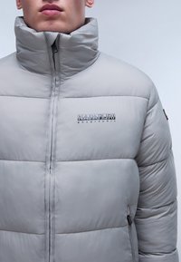 Grå pufferjacka med hög krage, med horisontellt kviltade sektioner, fickor med dragkedja och en logotyp på bröstet i kontrasterande svart.
