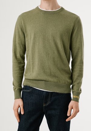 Mand iført olivengrøn crewneck sweater med ribbede manchetter og kant, kombineret med mørkeblå jeans mod en ensfarvet baggrund.