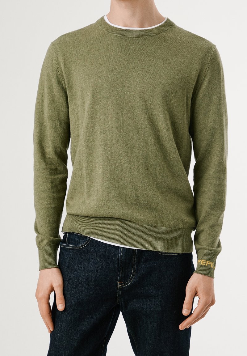 Homme portant un pull col rond vert olive avec poignets et ourlet côtelés, associé à un jean bleu foncé sur un fond uni.