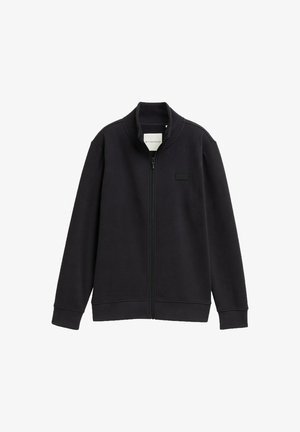 Musta vetskari, jossa on korkea kaulus, valmistettu pehmeästä kankaasta. Ominaisuuksiin kuuluu minimalistinen muotoilu ja pieni logopaneeli vasemmassa rinnassa.