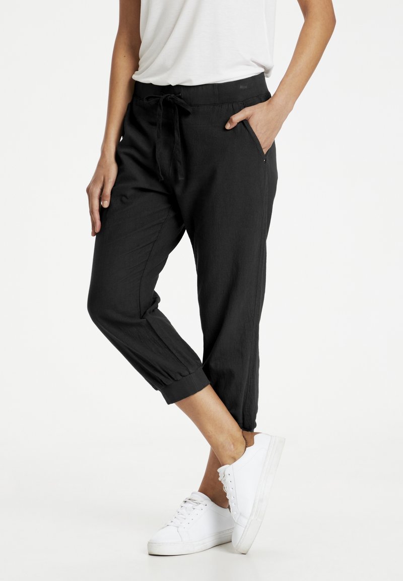 Pantalons noirs, légers et courts avec une taille élastique et un cordon de serrage, dotés de poches latérales et d'ourlets resserrés. Portés avec des baskets blanches.