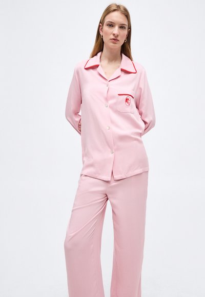 Γυναίκα στέκεται με τα χέρια στην πλάτη, φορώντας ανοιχτό ροζ σετ πιτζάμα από μετάξι με κόκκινες λεπτομέρειες και κεντημένη τσέπη, πάνω σε λευκό φόντο.