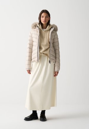 Světlý béžový péřový kabát s kapucí lemovanou kožešinou, nošený přes béžovou mikinu s kapucí a krémovou sukni. Obuv tvoří černé, strukturované boty.