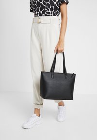 Sac fourre-tout synthétique noir avec deux poignées, une forme structurée et une fermeture éclair, tenu par une personne portant un pantalon de couleur neutre et des baskets.