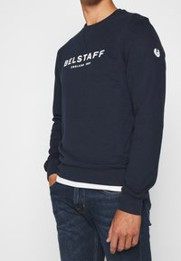 Mörkblå sweatshirt i bomull med rundad hals, ribbade ärmar och en vit logotryck på bröstet och vänster ärm.