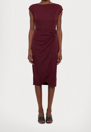 Kobieta ubrana w bordową sukienkę bez rękawów do kolan z marszczeniem w talii, w zestawieniu z czarnymi sandałami na obcasie z odkrytymi palcami, stojąca na jednolitym tle.