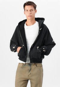 Black zip-up hoodie with a textured finish, featuring a lined hood, large pockets, and patterned cuffs. Worn over a white t-shirt.