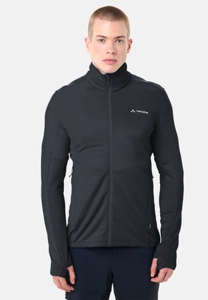 Junger Mann trägt eine schwarze Jacke mit Reißverschluss, Daumenlöchern und einem hohen Kragen, steht vor einem schlichten hellen Hintergrund.