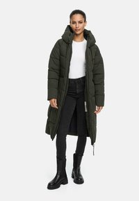 Manteau long en olive vert, rembourré avec une capuche, design matelassé et fermeture éclair à l'avant. Porté sur une chemise blanche et assorti avec un jean noir et des bottes.