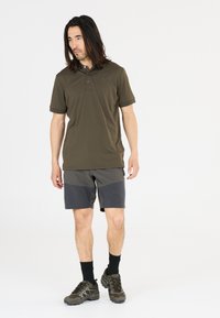 Oliviain vihreä polo-paita, jossa on kolmen napin lista, yhdistettynä harmaisiin shortsit, joissa on kulmatyylinen värilohko. Mustat sukat ja kengät viimeistelevät asun.