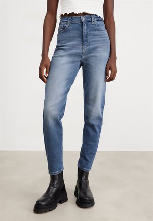 Persona con vaqueros azules de talle alto, top blanco corto y botas negras gruesas de tobillo, de pie sobre un suelo neutro con un fondo liso.