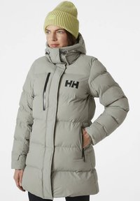 Šedá prešívaná bunda s vysokým golierom, čiernym logom, vreckami na zips a voľným strihom. Nosená so svetlozelenou čiapkou.