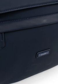 Borsa blu navy con una texture liscia, caratterizzata da una forma rettangolare e una targhetta con il logo. Include una patta pieghevole e dettagli cuciti.