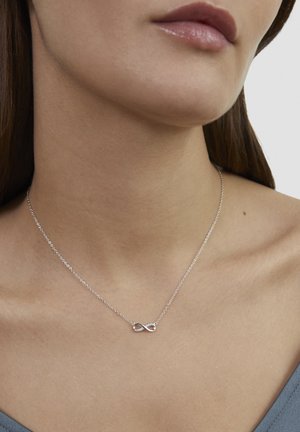 Silbernes Unendlichkeits-Halskette mit einer filigranen Kette; hat dezente Glitzereffekte in der Schleife, getragen an einem nackten Hals vor einem grauen Oberteil.