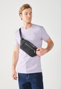 Black faux leather crossbody bag with a zip closure, featuring a textured pattern and an adjustable strap, worn by a model in a lavender shirt.