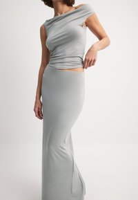 Helles graues, figurbetontes Kleid mit asymmetrischem Trägerdesign, fließendem Stoff und seitlichem Schlitz. Glatte Textur, schlichte und elegante Silhouette.