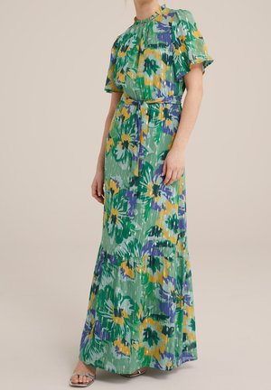 Vrouw draagt een groene maxi-jurk met een bloemenprint in geel, paars en wit, pofmouwen, een gerimpelde halslijn en een riem die in de taille gestrikt wordt.