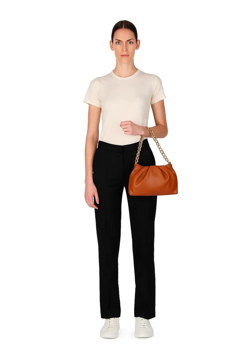 Donna in piedi, indossa una maglietta crema, pantaloni neri e sneakers bianche, mentre tiene una borsa in pelle marrone con una tracolla in catena d'oro sul braccio.