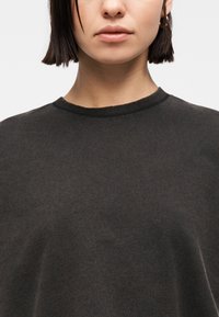 Γυναίκα με κοντά σκούρα μαλλιά που φορά μικρά χρυσά σκουλαρίκια κρίκους και σκούρο πουλόβερ με στρογγυλή λαιμόκοψη, σε απλό λευκό φόντο.