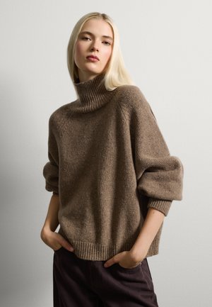Suéter marrón de punto con cuello alto, con dobladillo acanalado y mangas oversized, que presenta un acabado texturizado y un corte relajado. Llevado con pantalones oscuros.