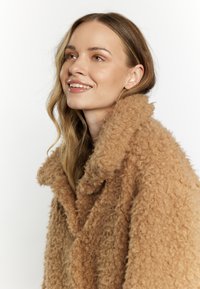 Fuzzy, lysebrun frakke med en stor krave, der har et struktureret stof. Yderbeklædningen har en løs pasform og et blødt udseende.