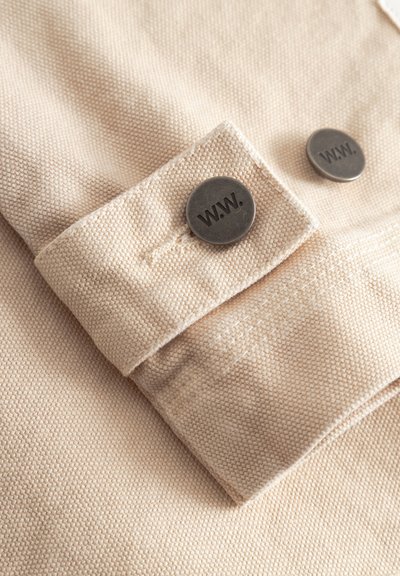 Beżowa tkanina o fakturze z ciemnymi okrągłymi metalowymi guzikami z wytłoczonym napisem "W.W." na złożonym mankiecie.