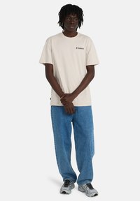 Μπεζ βαμβακερό μπλουζάκι με κοντά μανίκια και μικρό μαύρο λογότυπο στο στήθος. Συνδυασμένο με άνετο μπλε τζιν παντελόνι και γκρι αθλητικά παπούτσια.