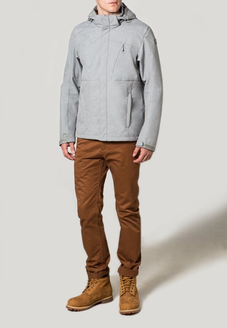 Homme portant une veste à capuche gris clair, un pantalon marron et des bottes lacées couleur beige, debout devant un fond uni.