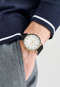 Orologio tondo con cassa dorata, quadrante bianco, indici numerici scuri e cinturino in pelle nera, indossato su un polso con tessuto grigio.
