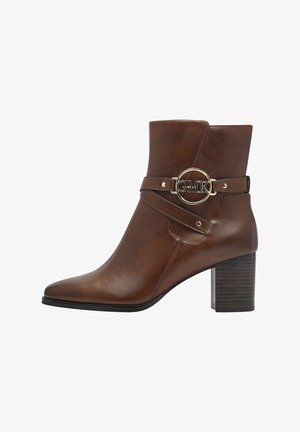Brown leather ankle boot with a pointed toe, stacked heel, and gold circular hardware featuring "GMK" on the strap. Textured finish.