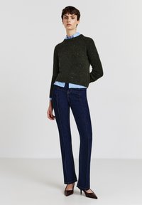 Dunkelgrüner Strickpullover mit rundem Ausschnitt, über einem hellblauen Hemd getragen. Kombiniert mit dunkelblauen Schlagjeans und braunen Absatzschuhen.