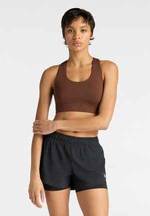 Jonge vrouw met kort haar, gekleed in een bruine sportbeha en zwarte sportshorts, staand met één arm gekruist over haar lichaam.