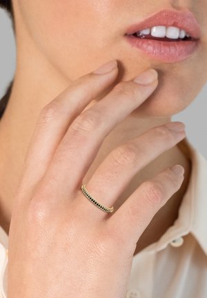 Main avec une fine bague en or ornée de petites pierres noires près des lèvres, sur fond neutre et vêtement de couleur claire.