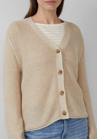 Beżowy sweter zapinany na guziki z dekoltem w serek, z czterema drewnianymi guzikami i teksturowanym materiałem. Nosi się go na paskowaną bluzkę. Swobodny krój.