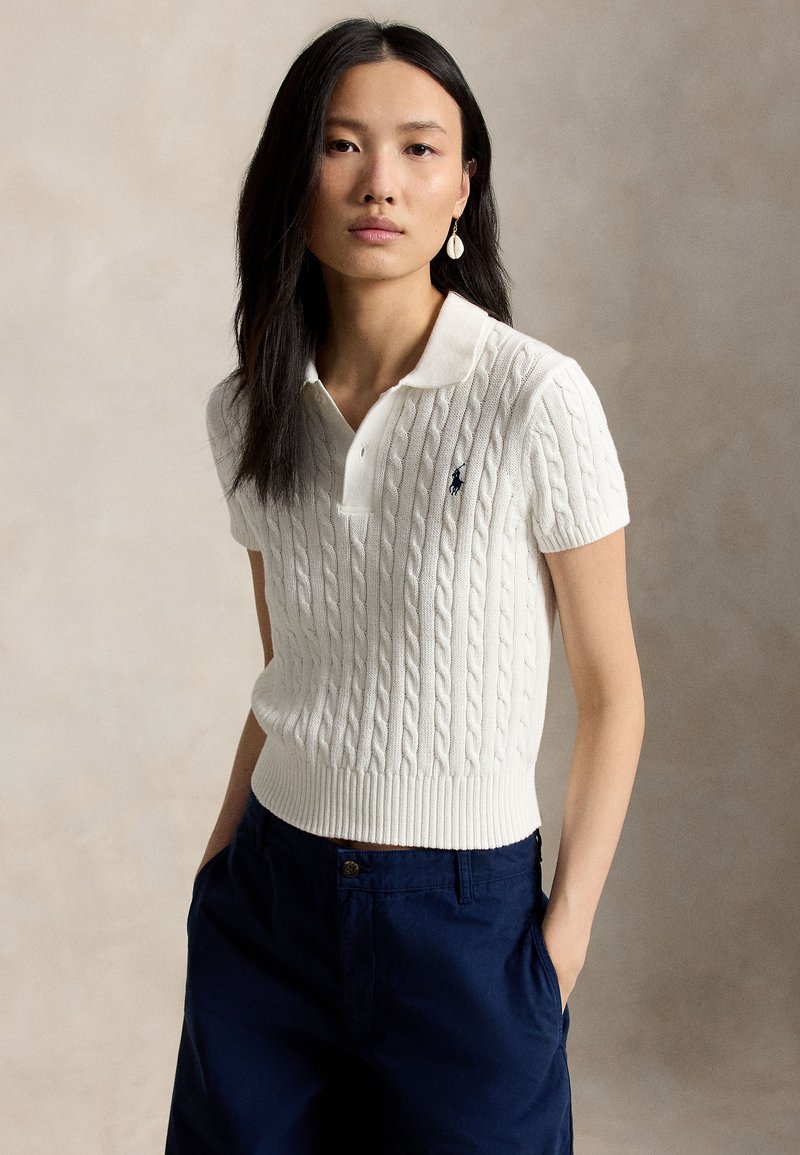 Woman with long dark hair wears white short-sleeve cable-knit polo shirt and navy pants, standing with hands in pockets against plain background.